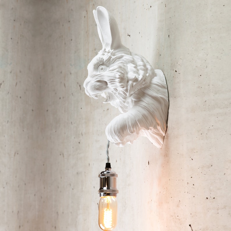 May include: White ceramic rabbit wall sconce with a hanging light bulb. The rabbit's head is turned to the side and its ears are perked up. The light bulb is a clear glass bulb with a silver metal socket.