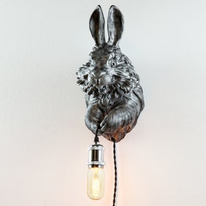 May include: A silver rabbit wall sconce with a black cord and a light bulb. The rabbit is holding the cord in its paw.