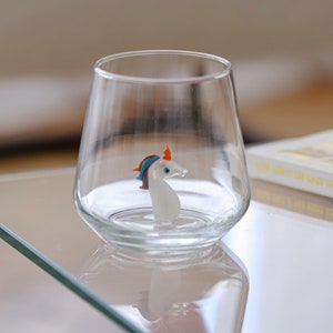 May include: A clear glass with a white unicorn figurine inside, sitting on a glass table top. Two red tulips with green stems are in the background.