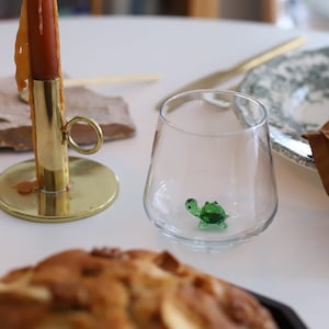 May include: A lit candle in a brass holder sits on a white table next to a clear glass with a green glass turtle inside. A brown napkin and a plate with a green and white pattern are in the background.