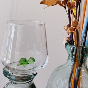 May include: A clear glass bottle with a green glass top. The top is shaped like a wine glass and has a small green glass turtle inside. The bottle is sitting on a table next to a clear glass vase with brown dried flowers and colorful glass straws.