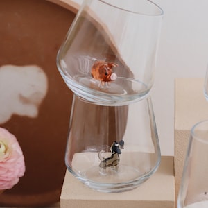 May include: Two clear glass cups with a small pig figurine in the top cup and a small dog figurine in the bottom cup. The cups are stacked on top of each other and are sitting on a beige surface.