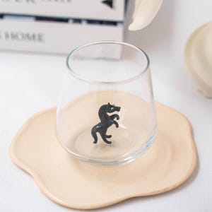 May include: A clear glass cup with a black horse figurine inside, sitting on a light beige saucer.