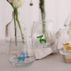 May include: Four clear glass tumblers with colorful glass dog figurines inside. The tumblers are sitting on a white surface.