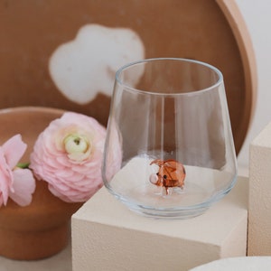 May include: A clear glass tumbler with a small brown glass pig figurine inside. The glass is sitting on a beige square pedestal.