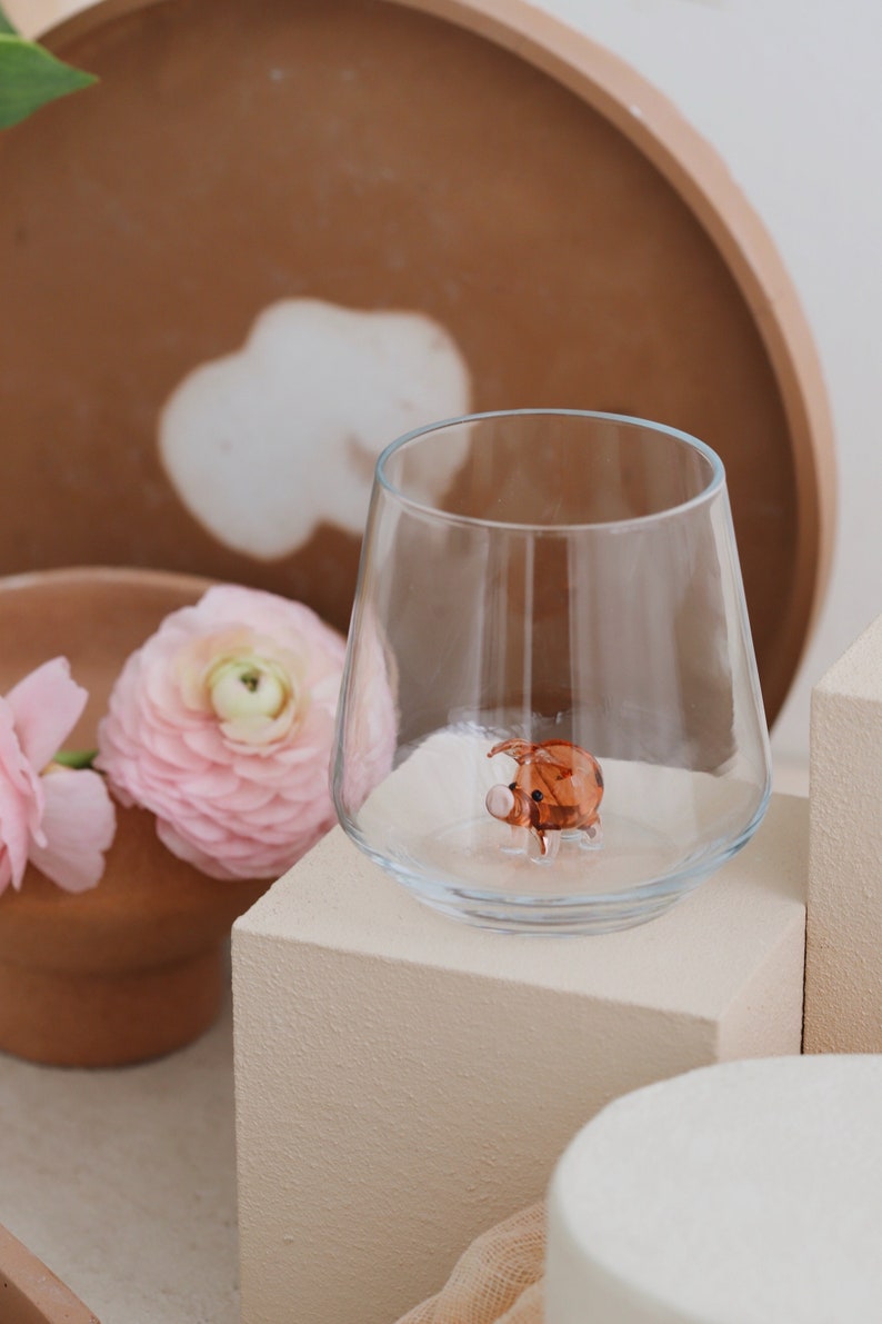 May include: A clear glass tumbler with a small brown glass pig figurine inside. The glass is sitting on a beige square pedestal.