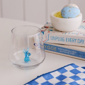 May include: A blue glass rabbit figurine sits inside a clear glass tumbler. Two books with colourful covers are stacked on top of each other. The top book has the text "Unplug Every Day" on the cover. The bottom book has the text "Ygge Danimarkalilarin Mutluluk Sirri Meik Wik" on the cover. A white bowl with blue and yellow speckled Easter eggs sits on top of the books.