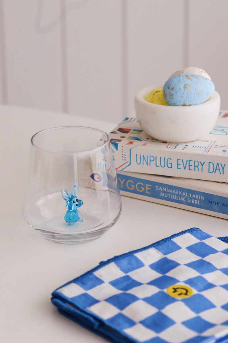 May include: A blue glass rabbit figurine sits inside a clear glass tumbler. Two books with colourful covers are stacked on top of each other. The top book has the text "Unplug Every Day" on the cover. The bottom book has the text "Ygge Danimarkalilarin Mutluluk Sirri Meik Wik" on the cover. A white bowl with blue and yellow speckled Easter eggs sits on top of the books.