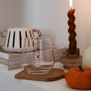 May include: A clear glass with a small white ghost figurine inside, sitting on a wooden coaster. A lit candle, a pumpkin, and a white ceramic vase with black stripes are in the background.