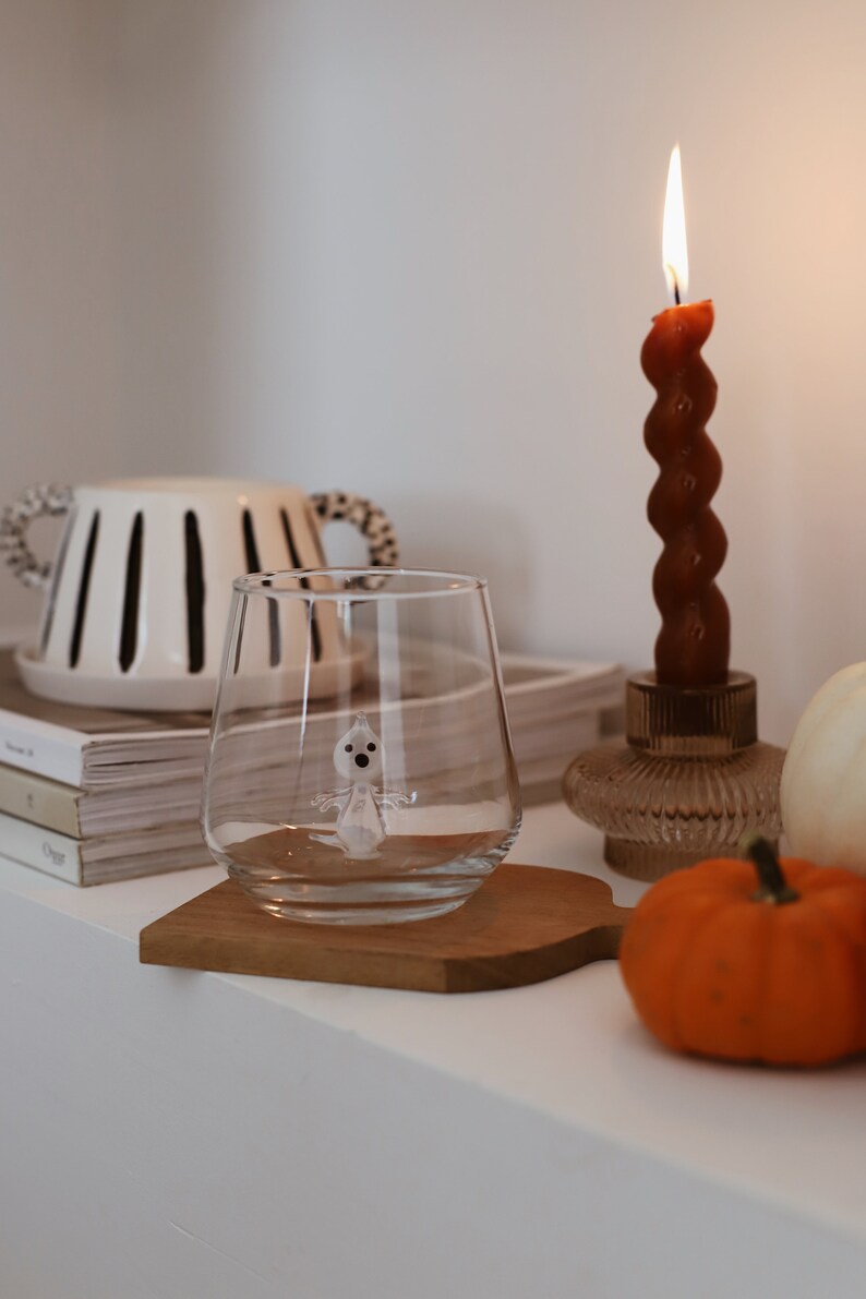 May include: A clear glass with a small white ghost figurine inside, sitting on a wooden coaster. A lit candle, a pumpkin, and a white ceramic vase with black stripes are in the background.