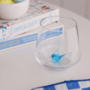 May include: A clear glass with a blue glass bird figurine inside. The glass is sitting on top of a stack of books with the text "UNPLUG EVERY DAY" and "HYGGE" visible. A blue and white checked cloth is in the foreground.