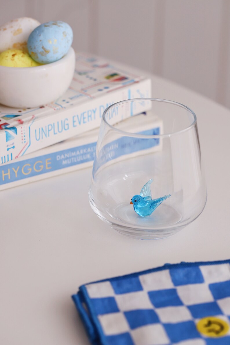 May include: A clear glass with a blue glass bird figurine inside. The glass is sitting on top of a stack of books with the text "UNPLUG EVERY DAY" and "HYGGE" visible. A blue and white checked cloth is in the foreground.