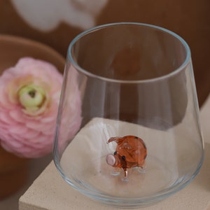 May include: A small, clear glass with a brown glass pig figurine inside. The pig has a pink snout and black eyes.