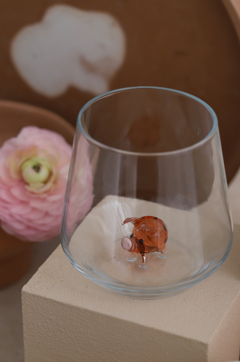 May include: A small, clear glass with a brown glass pig figurine inside. The pig has a pink snout and black eyes.