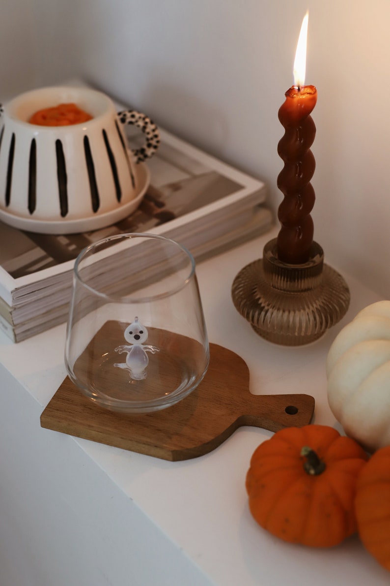 May include: A white ceramic candle holder with black stripes and a small ghost figurine inside a clear glass. The candle holder is on a stack of magazines next to a brown candle in a glass holder, a white pumpkin, and two orange pumpkins.