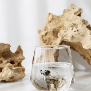 May include: A clear glass filled with water and a small black and white dog figurine inside. The glass is sitting on a white surface with a light brown rock behind it.