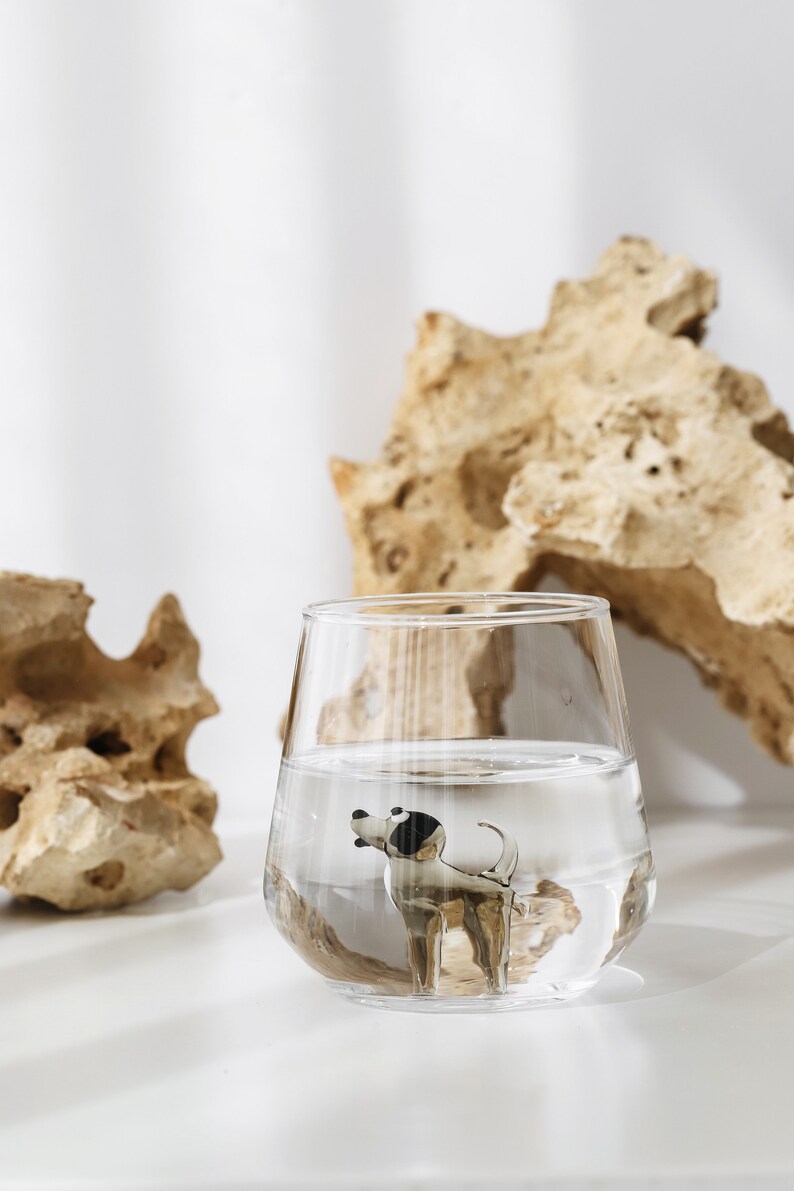 May include: A clear glass filled with water and a small black and white dog figurine inside. The glass is sitting on a white surface with a light brown rock behind it.
