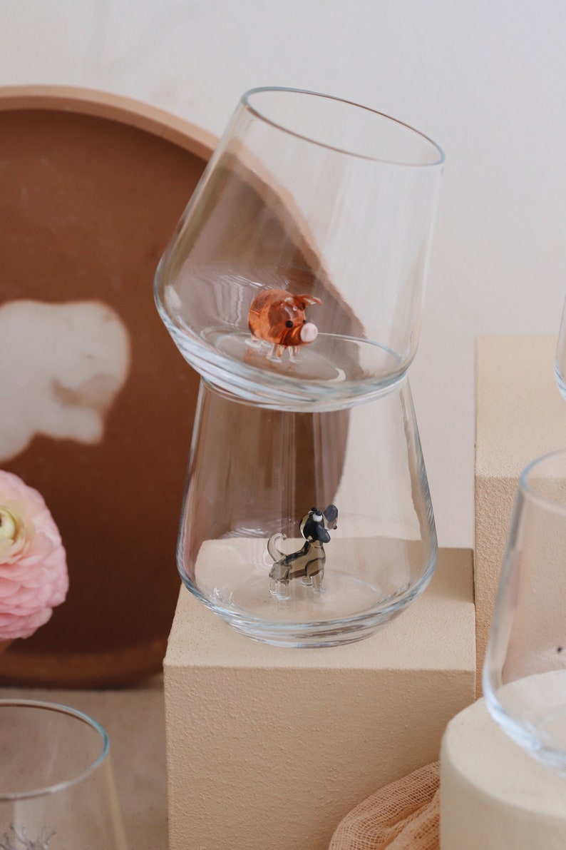 May include: Two clear glass cups with a small pig figurine in the top cup and a small dog figurine in the bottom cup. The cups are stacked on top of each other and are sitting on a beige surface.