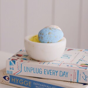 May include: Two books stacked on top of each other. The top book is titled "Unplug Every Day" and the bottom book is titled "Hygge:  DanimarKalilarin Mutluluk Sirri" by Meik Wiking. A white bowl with three blue and yellow speckled Easter eggs sits on top of the books.