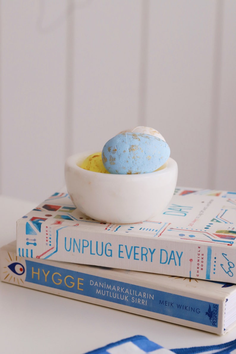 May include: Two books stacked on top of each other. The top book is titled "Unplug Every Day" and the bottom book is titled "Hygge:  DanimarKalilarin Mutluluk Sirri" by Meik Wiking. A white bowl with three blue and yellow speckled Easter eggs sits on top of the books.