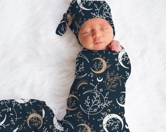 Könnte beinhalten: Ein neugeborenes Baby, eingewickelt in einen dunkelblauen Swaddle mit einem weißen und goldenen Halbmond- und Blumenmuster. Das Baby trägt eine passende dunkelblaue Mütze mit dem gleichen Muster.