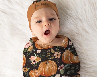 May include: A baby swaddled in a black and brown swaddle with a pumpkin and floral pattern. The baby is wearing a brown hat with the name "Jeremiah" written on it.