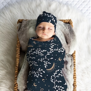 May include: A sleeping baby swaddled in a navy blue swaddle with a white and gold leaf and crescent moon pattern. The baby is wearing a matching navy blue hat with the same pattern.