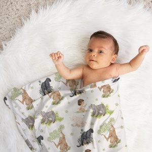 Op de afbeelding: Een baby gewikkeld in een witte muslin swaddle deken met een jungle dierenprint. De deken heeft zwarte panters, bruine beren en een junglesc&egrave;ne.