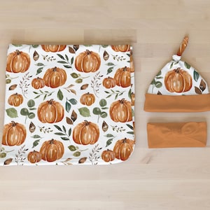 May include: A set of three baby items featuring a watercolor pumpkin and leaf pattern. The set includes a blanket, a hat, and a headband. All items are in shades of orange and brown.