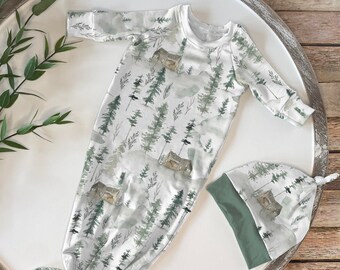 Op de afbeelding: Een witte babyjurk met een groene en witte aquarelprint van bomen en huizen. De jurk heeft lange mouwen en een geknoopte onderkant. Een bijpassende groene en witte muts is inbegrepen.