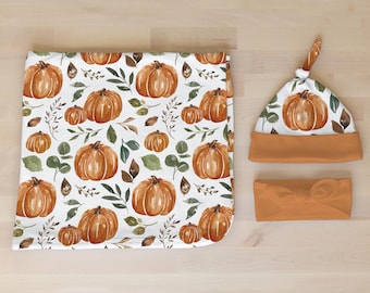 May include: A set of three baby items featuring a watercolor pumpkin and leaf pattern. The set includes a blanket, a hat, and a headband. All items are in shades of orange and brown.