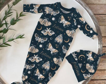 May include: A dark teal baby gown and matching hat set. The set features a repeating pattern of moths, moons, and stars in cream and gold. The name "Katrina" is printed on both items. The gown has long sleeves and a knotted bottom.