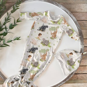 Op de afbeelding: Een witte babyjurk met een jungledierenprint met een tijger, panter, beer en een jongensfiguur. De jurk heeft een geknoopte staart en wordt geleverd met een bijpassende muts.