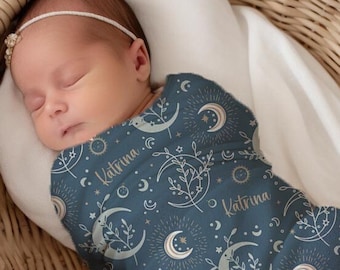 May include: A baby swaddled in a blue and gold swaddle with a moon and stars pattern. The swaddle has the name "Katrina" printed on it.