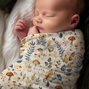 May include: A baby swaddled in a white muslin swaddle with a watercolor pattern of leaves, mushrooms, and acorns. The swaddle is a popular choice for newborns and infants.