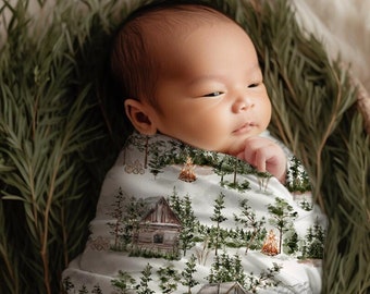 Op de afbeelding: Een pasgeboren baby gewikkeld in een witte mousseline-wikkeldeken met een print van een boshuisje en kampvuur. De baby slaapt rustig in een gevlochten mand gevuld met groen gebladerte.