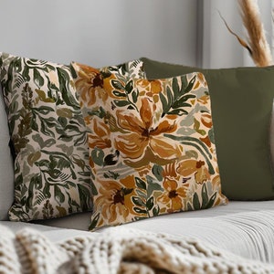 Op de afbeelding: Twee kussens met bloemenpatronen. Een kussen heeft een groen en wit bloemenpatroon, en de andere heeft een geel en groen bloemenpatroon. De kussens liggen op een witte bank met een cr&egrave;mekleurige gebreide deken.