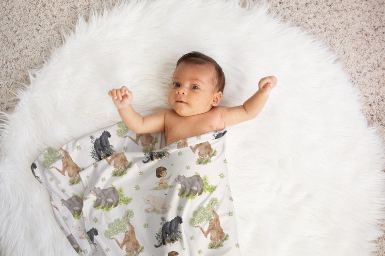 Op de afbeelding: Een baby gewikkeld in een witte muslin swaddle deken met een jungle dierenprint. De deken heeft zwarte panters, bruine beren en een junglesc&egrave;ne.