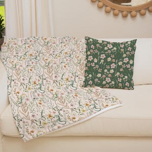 May include: A white throw blanket with a floral and snake pattern is draped over a white couch. A green throw pillow with a floral pattern is on the couch.
