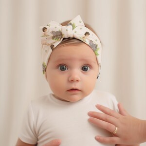 Op de afbeelding: Een baby die een wit hoofdband draagt met een print van een cartoonfiguur en dieren. Het hoofdband is bovenop het hoofd van de baby in een strik geknoopt.