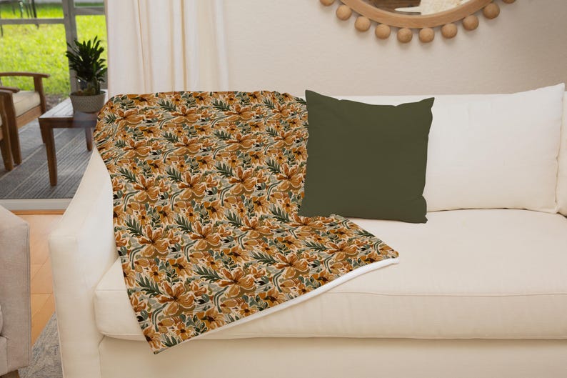 Op de afbeelding: Een deken met een bloemenpatroon in tinten geel, oranje en groen. De deken is gevouwen en gedrapeerd over een witte bank met een donkergroen kussen.