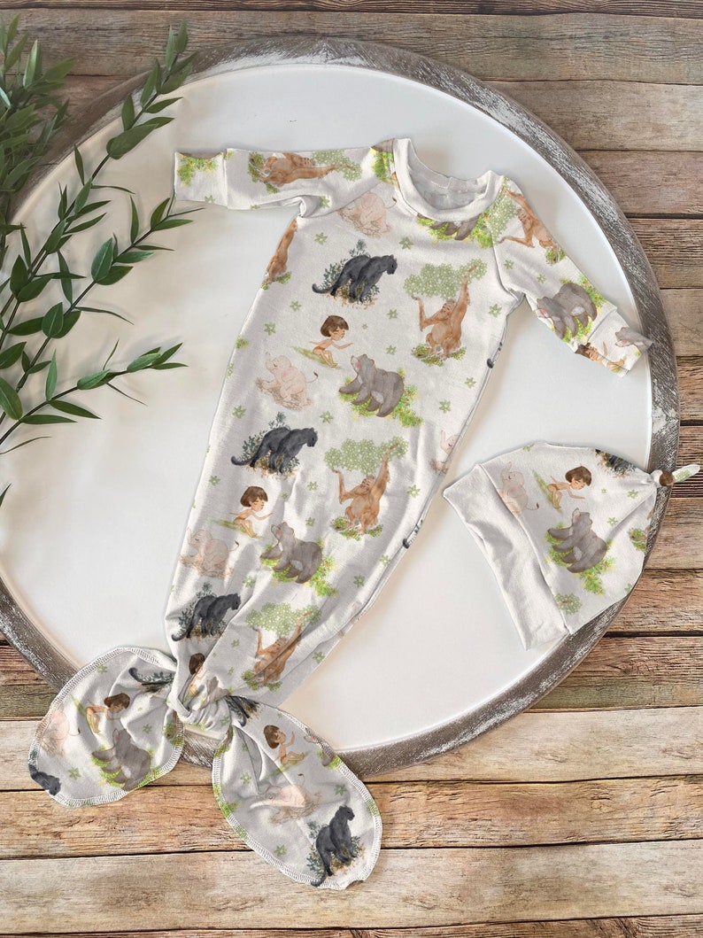 Op de afbeelding: Een witte babyjurk met een jungledierenprint met een tijger, panter, beer en een jongensfiguur. De jurk heeft een geknoopte staart en wordt geleverd met een bijpassende muts.