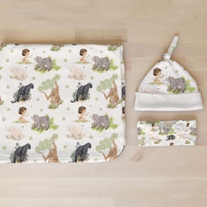 Op de afbeelding: Een witte babydeken, muts en hoofdband met een jungle thema. De stof heeft een herhalend patroon van een jongen, een panter, een beer en een olifant.