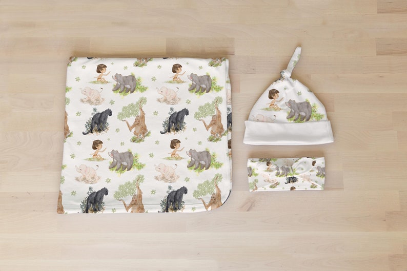 Op de afbeelding: Een witte babydeken, muts en hoofdband met een jungle thema. De stof heeft een herhalend patroon van een jongen, een panter, een beer en een olifant.