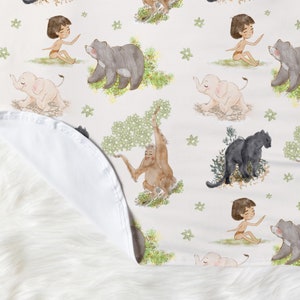 Op de afbeelding: Een witte babydeken van stof met een herhalend patroon van jungledieren, waaronder een olifant, een beer, een aap en een jongen. De dieren zijn ge&iuml;llustreerd in aquarelstijl met groen gebladerte.