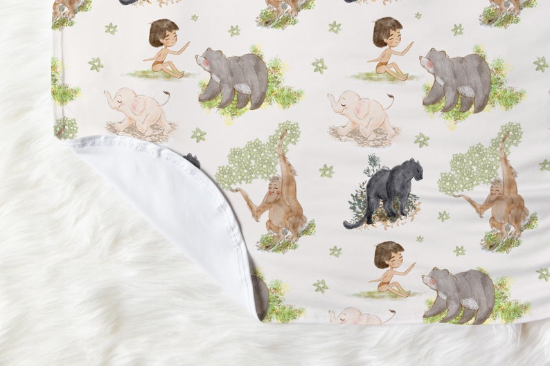 Op de afbeelding: Een witte babydeken van stof met een herhalend patroon van jungledieren, waaronder een olifant, een beer, een aap en een jongen. De dieren zijn ge&iuml;llustreerd in aquarelstijl met groen gebladerte.