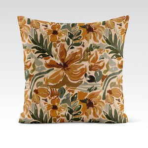Op de afbeelding: Een decoratief kussen met een bloemenpatroon met grote, abstracte bloemen in tinten oranje, geel en bruin. De achtergrond is cr&egrave;mekleurig met groene bladeren en takken.
