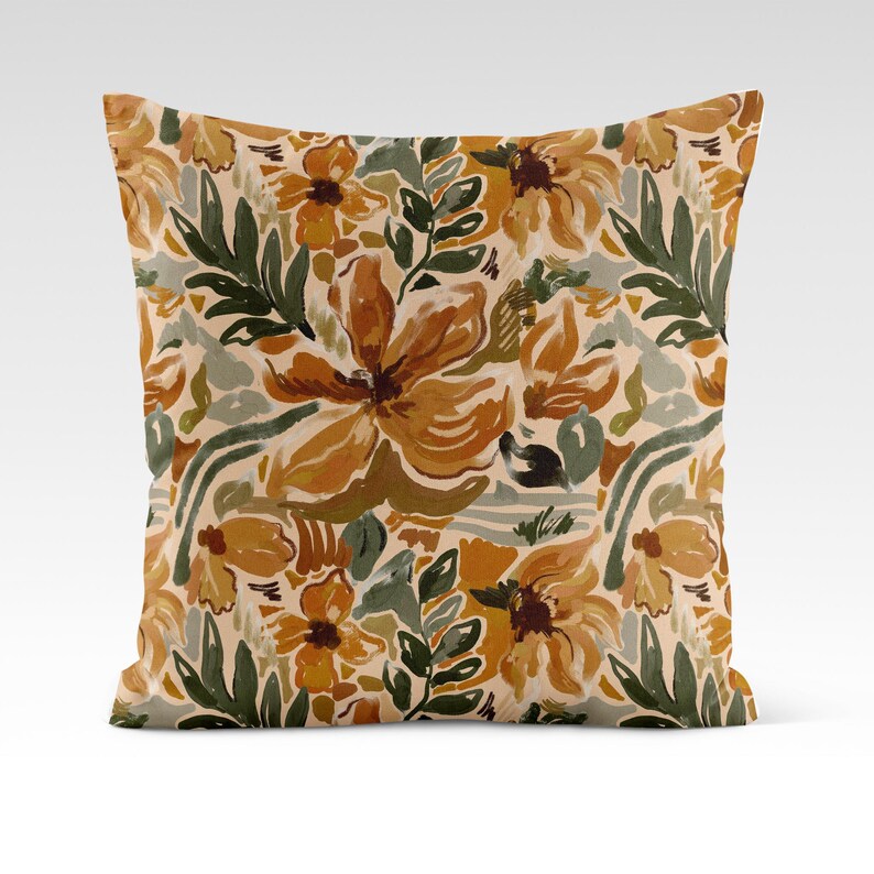 Op de afbeelding: Een decoratief kussen met een bloemenpatroon met grote, abstracte bloemen in tinten oranje, geel en bruin. De achtergrond is cr&egrave;mekleurig met groene bladeren en takken.