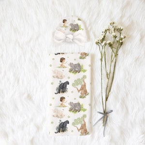 Op de afbeelding: Een wit babydekentje met een jungle thema met een jongetje, dieren en groene bladeren. Er is ook een bijpassende witte muts met strik.