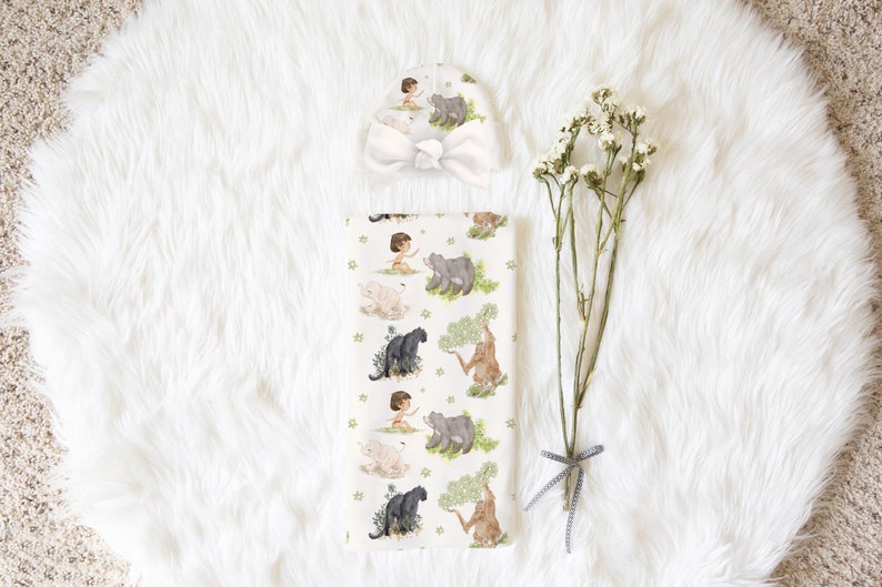 Op de afbeelding: Een wit babydekentje met een jungle thema met een jongetje, dieren en groene bladeren. Er is ook een bijpassende witte muts met strik.